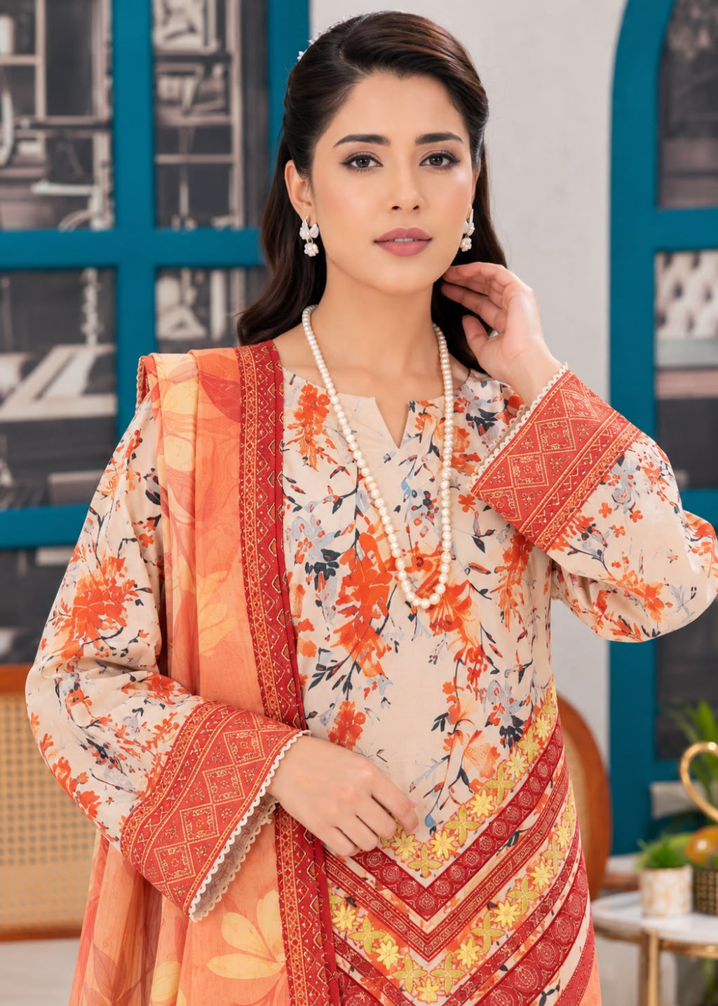 A woman models a traditional three-piece salwar kameez suit in a studio setting. The suit features a beige base with orange and red floral prints, paired with a matching orange dupatta and a red patterned border on the sleeves and kameez front.