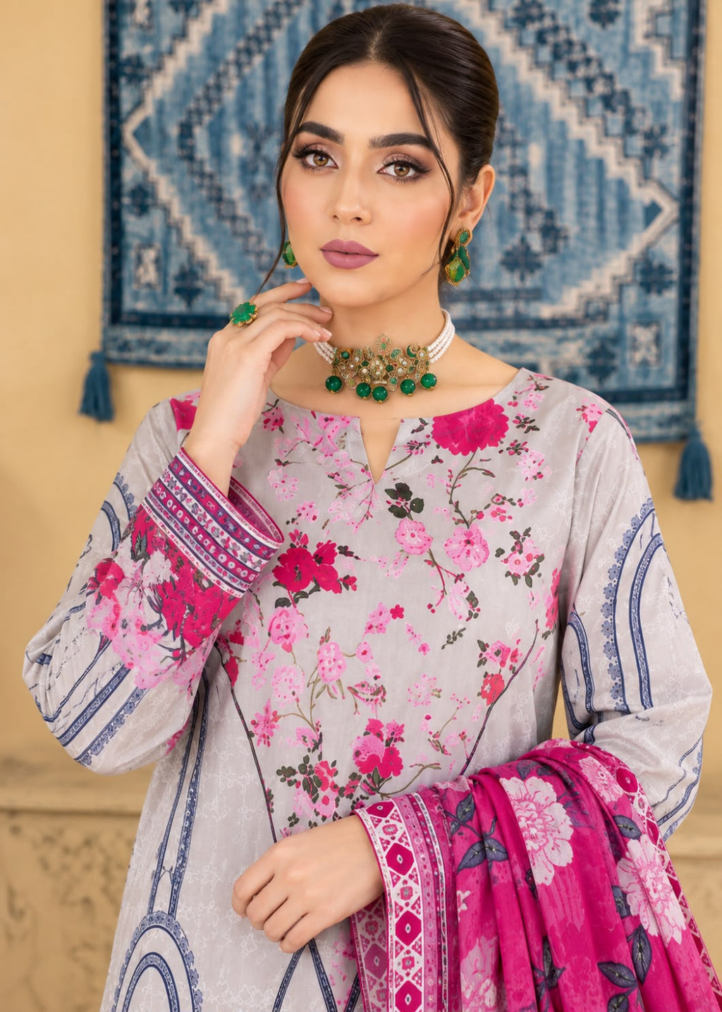 A woman models a light gray three-piece ethnic suit with pink and magenta floral prints, featuring a matching printed dupatta and green beaded jewelry against a warm beige wall with a blue patterned tapestry.
