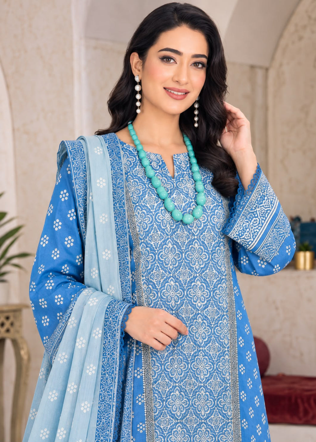 A woman modeling a blue printed traditional outfit with a matching dupatta, featuring intricate white and light blue patterns, a turquoise beaded necklace, and dangling earrings in an indoor setting.