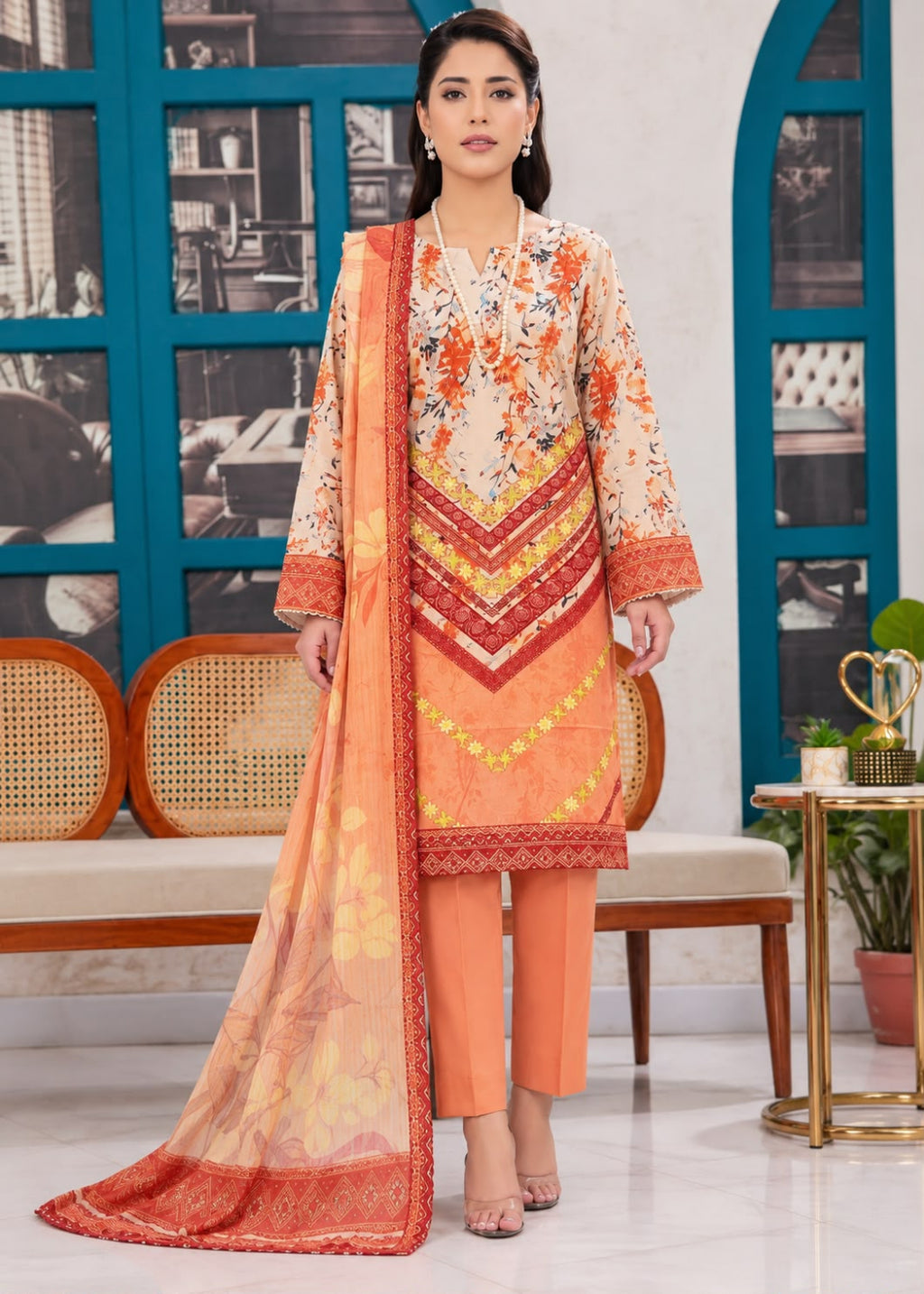 A woman models a traditional three-piece salwar kameez suit in a studio setting. The set features a beige kurta with orange, red, and yellow floral prints and chevron embroidery, paired with matching orange trousers and a sheer orange dupatta with a red border.
