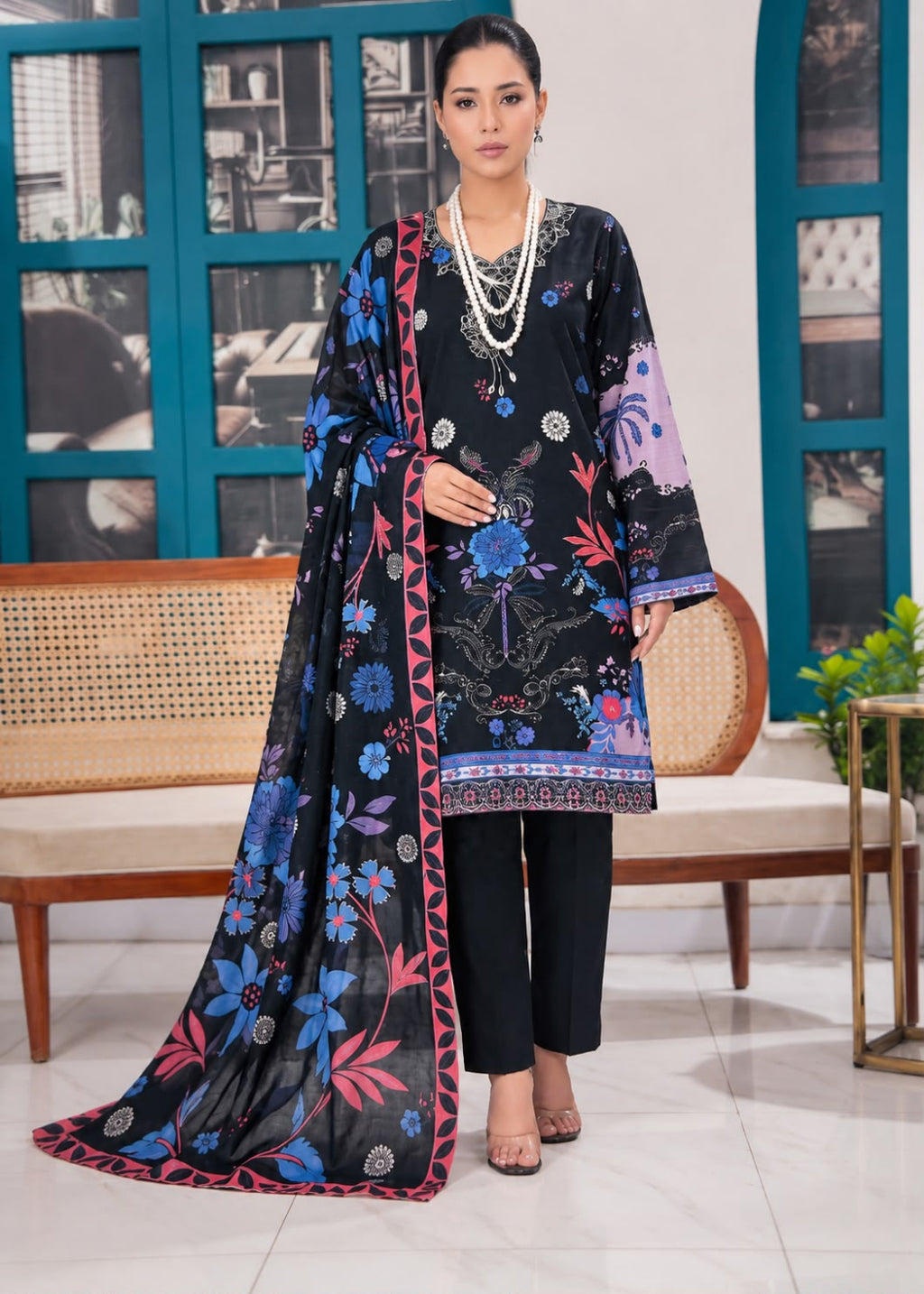 A woman models a black 3-piece printed salwar kameez suit with a matching dupatta. The suit features vibrant blue, pink, and purple floral patterns, paired with black trousers and accessorized with a multi-strand pearl necklace.