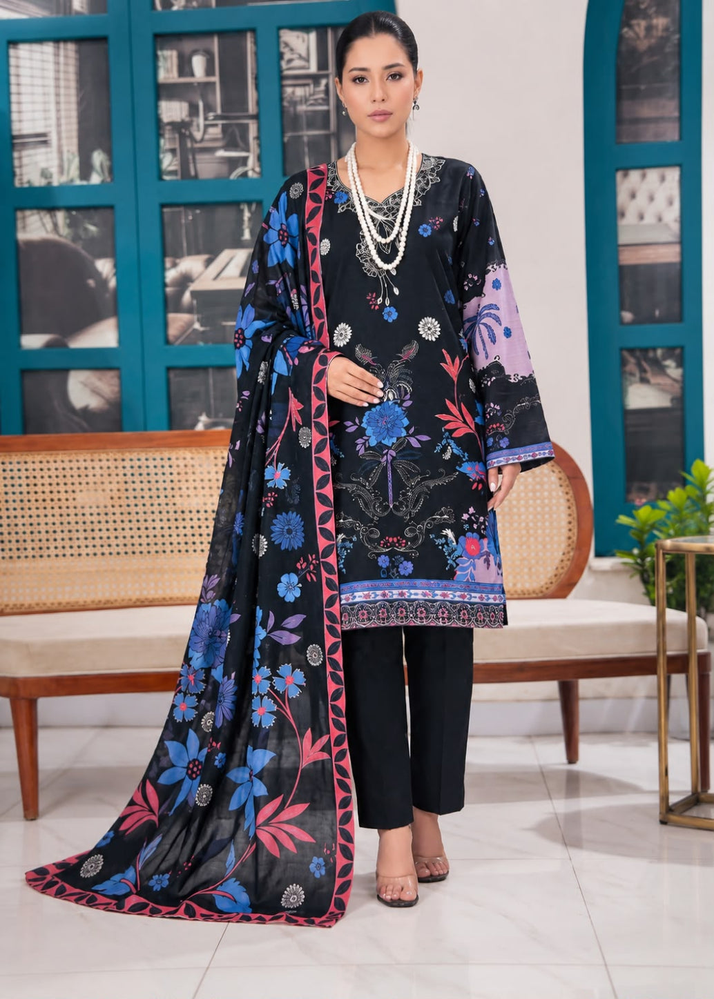 A woman models a black 3-piece printed salwar kameez suit with a matching dupatta. The suit features vibrant blue, pink, and purple floral patterns, paired with black trousers and accessorized with a multi-strand pearl necklace.