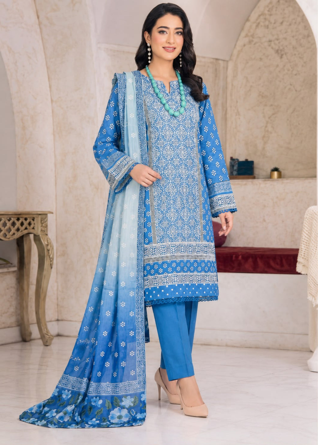 A woman models a traditional blue Indian salwar kameez suit set with a matching dupatta in a beautifully decorated indoor setting. The set features a blue printed kurta, solid blue trousers, and a blue patterned dupatta.