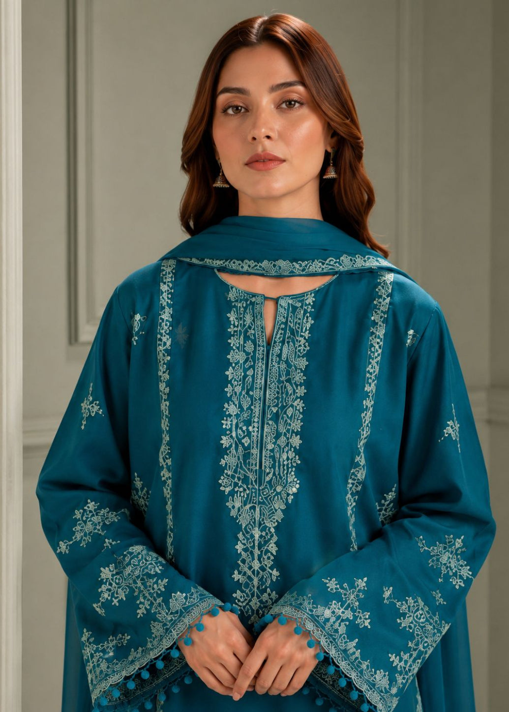 A woman wearing a teal blue embroidered traditional outfit with a matching dupatta, standing against a neutral paneled wall.