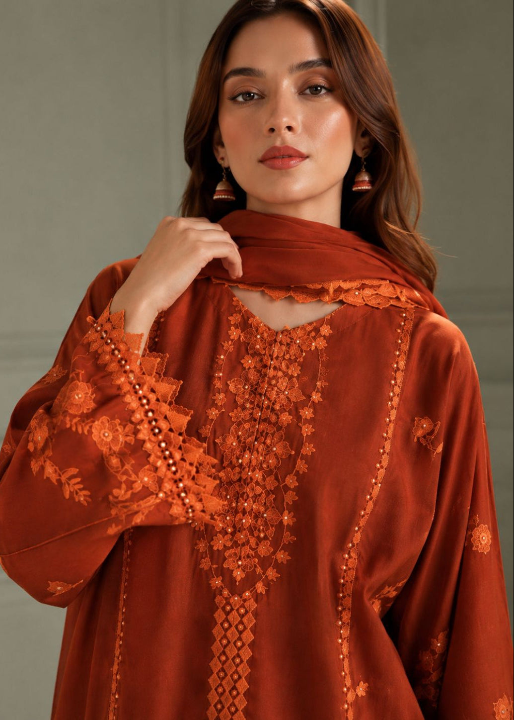 A woman wearing a rust-orange embroidered kurta with a matching dupatta, featuring intricate floral embroidery and beadwork details.