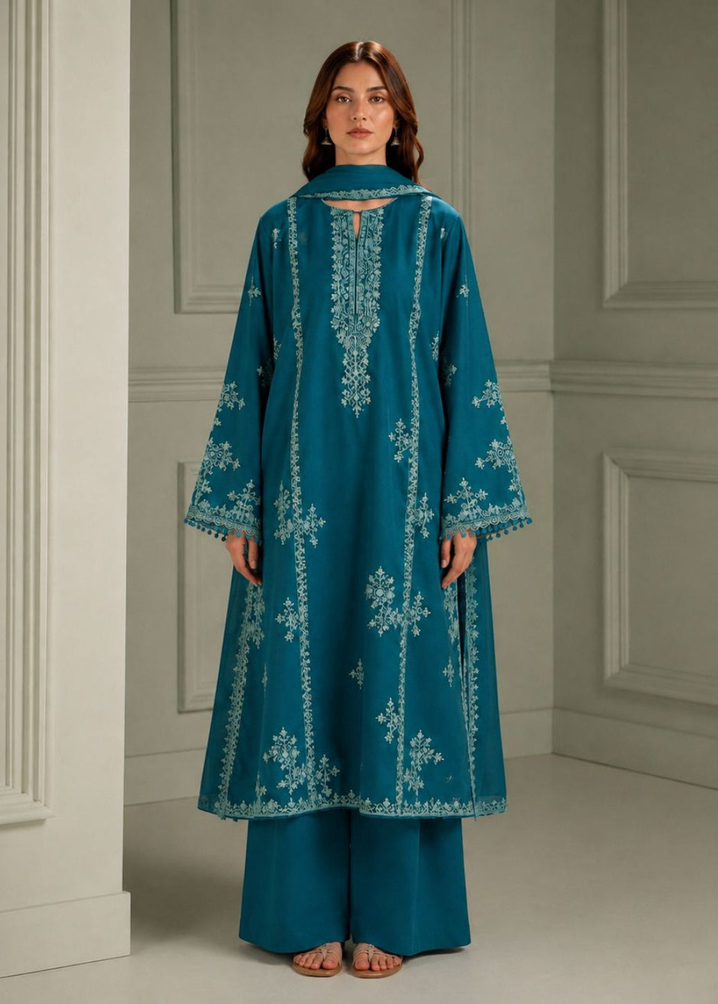 A woman stands indoors wearing a teal embroidered tunic with matching wide-leg pants and a coordinating scarf. The tunic has white floral embroidery and a keyhole cutout at the collar.