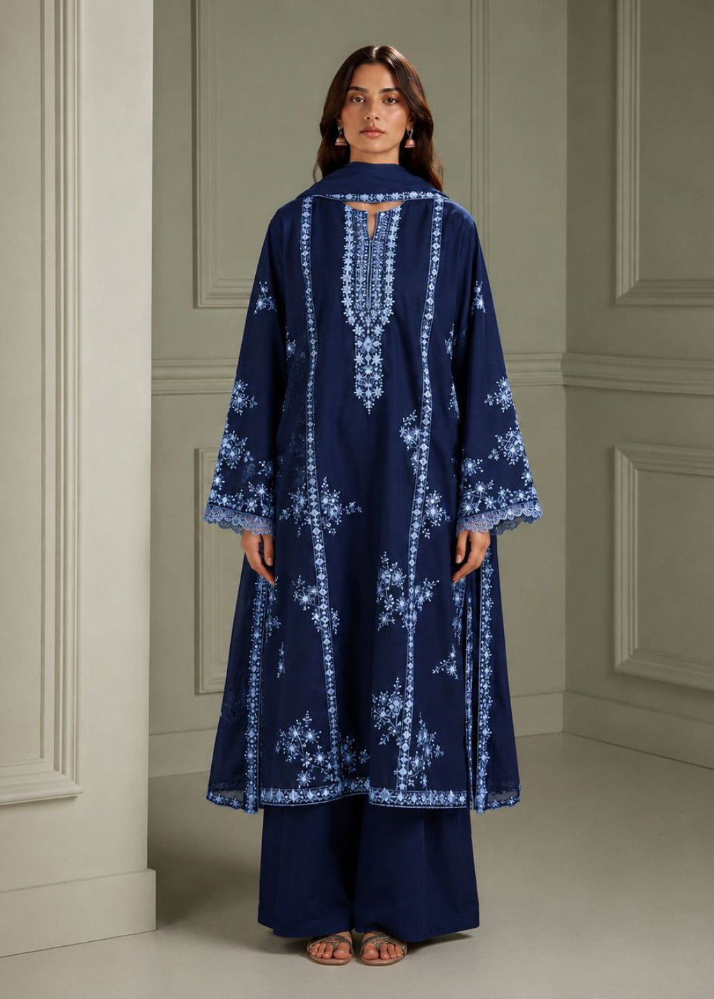A woman stands facing away from the camera wearing a navy blue long kurta with intricate white floral embroidery and matching navy blue wide-leg trousers in an elegant indoor setting with paneled walls.