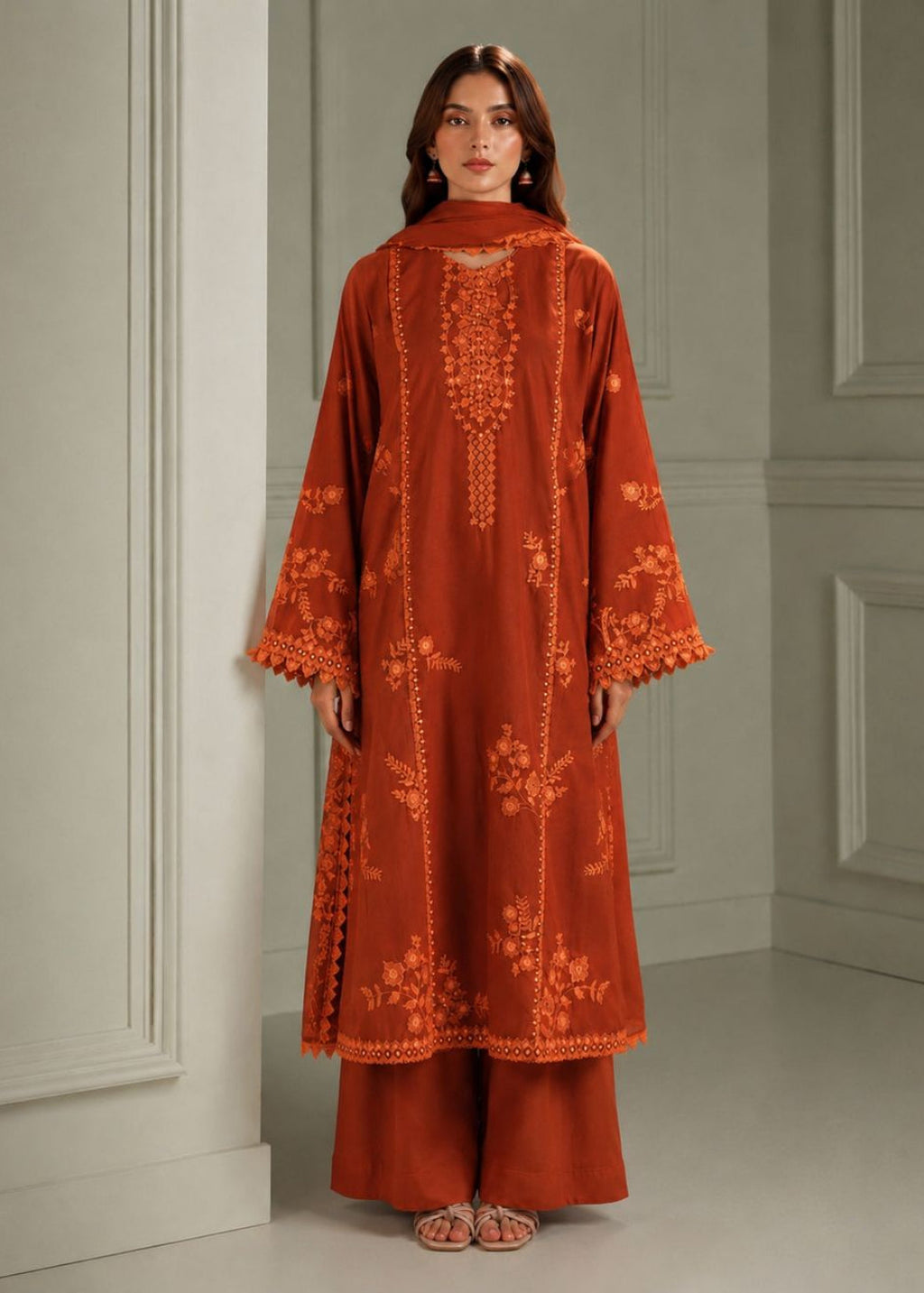 A woman stands indoors wearing a rust-orange embroidered kurta with matching wide-leg pants and a dupatta. The outfit has ornate floral embroidery and scalloped trim.