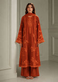 A woman stands indoors wearing a rust-orange embroidered kurta with matching wide-leg pants and a dupatta. The outfit has ornate floral embroidery and scalloped trim.