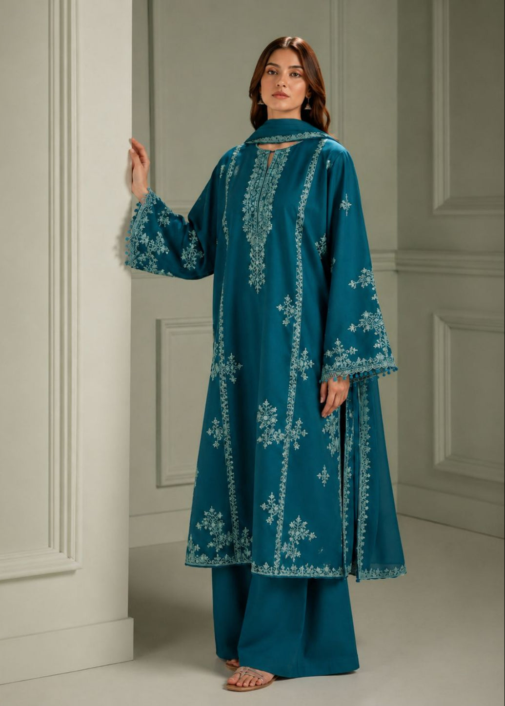 A woman models a teal blue embroidered kurta and matching wide-leg trousers in an elegant indoor setting with white paneled walls.