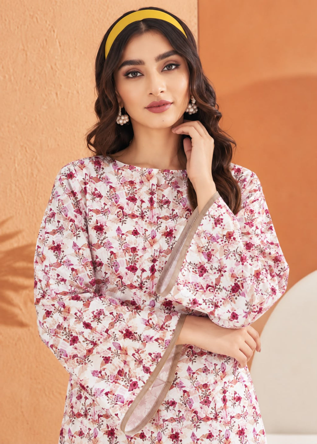A woman modeling a long-sleeved floral print kurta in pink and red tones on a white base, paired with a yellow headband and dangling earrings, set against a warm orange textured wall.