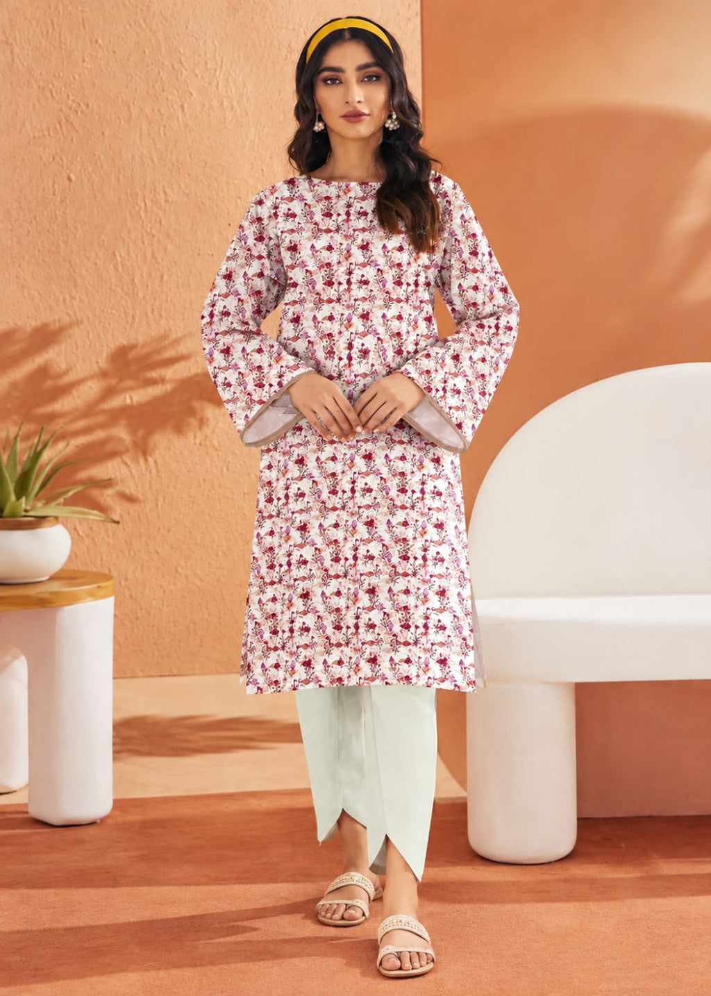 A woman models a long-sleeve kurta with a pink and white floral pattern, paired with light sage green pants and beige sandals, standing in a modern, warm-toned studio setting.