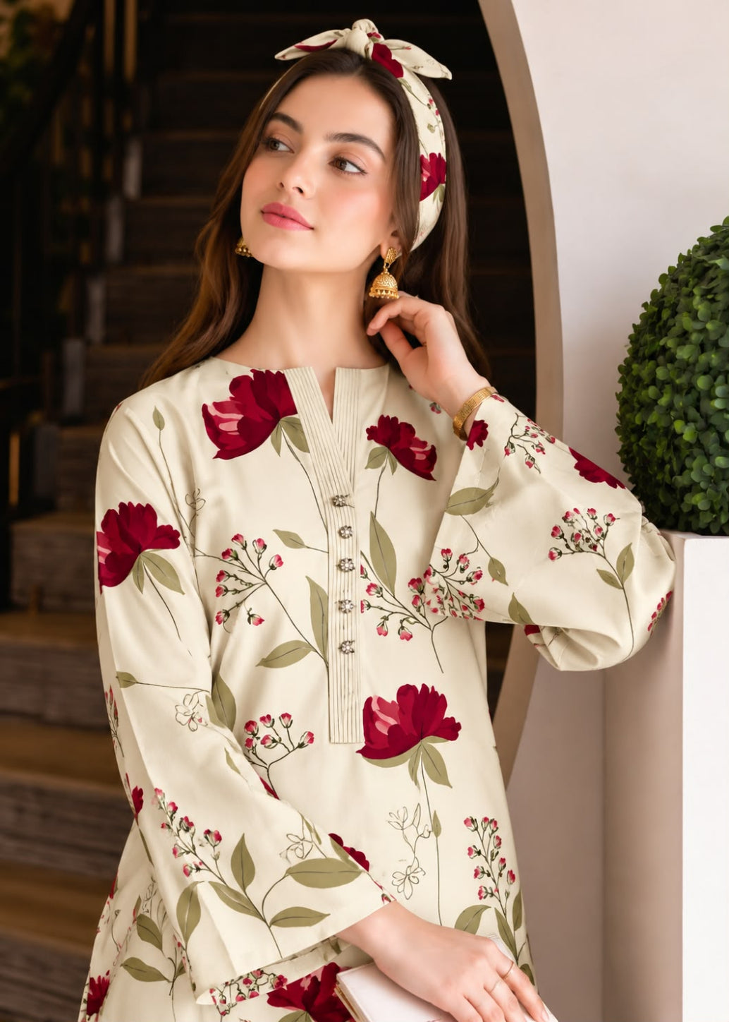 A woman wearing a cream-colored long-sleeve tunic dress with a red and green floral print, a matching cream headband, and gold earrings, posing in an elegant indoor setting.