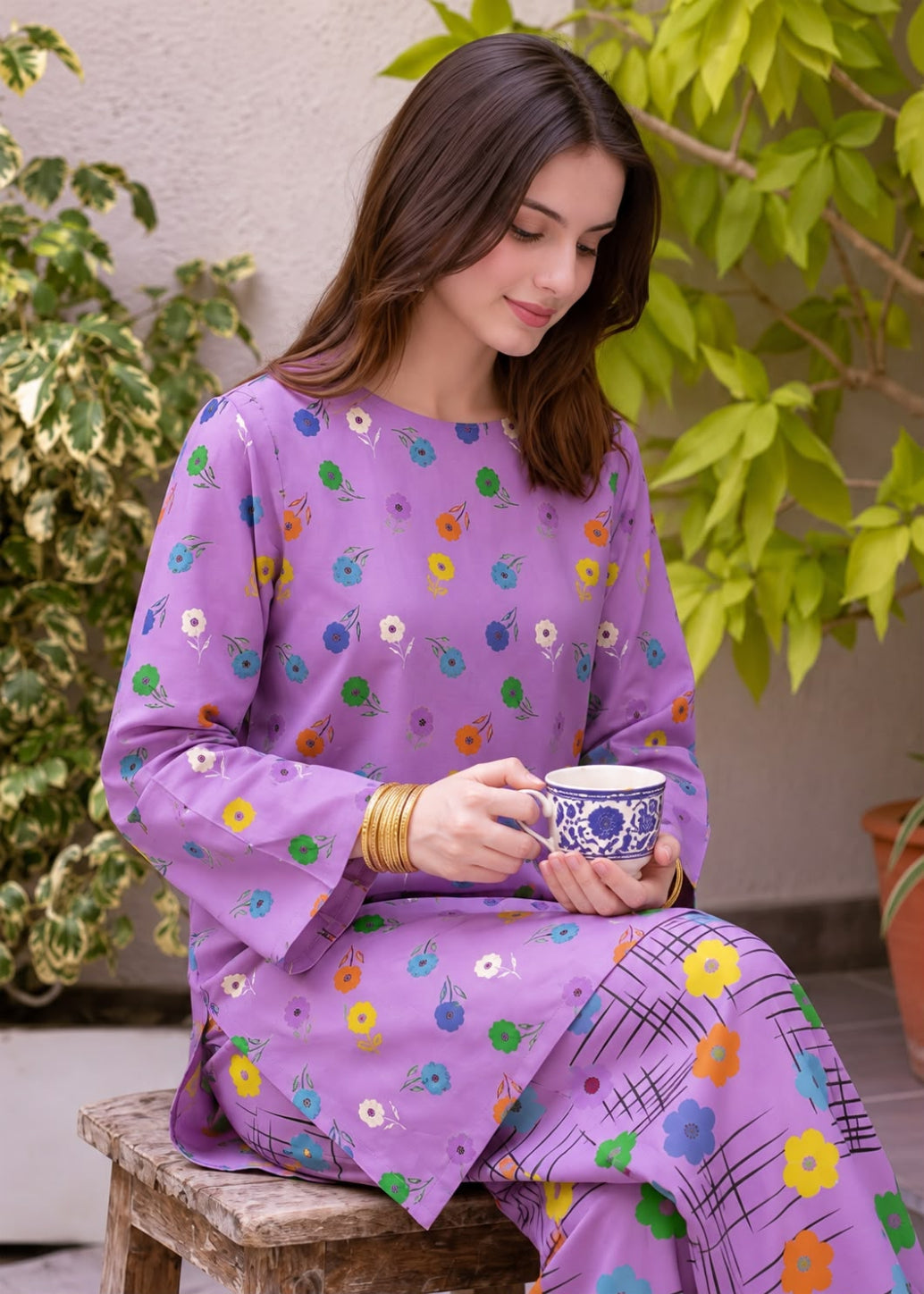 A woman wearing a purple long-sleeve kurta with colorful floral prints sits on a wooden stool outdoors, holding a blue and white patterned cup.