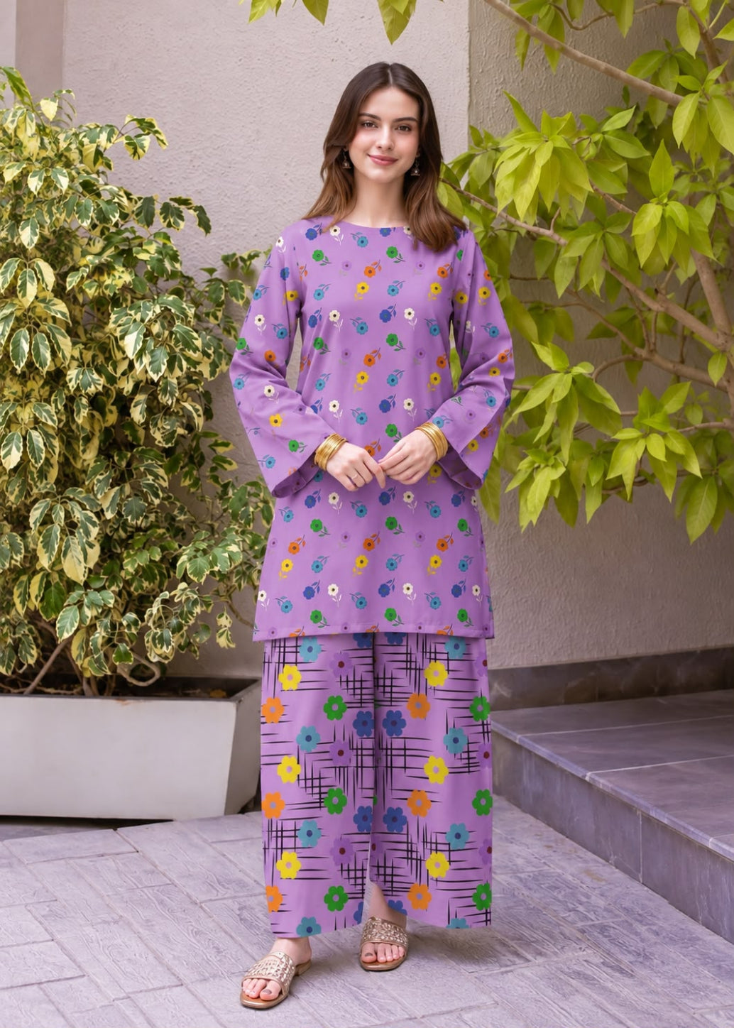 A woman modeling a purple two-piece kurta set with colorful floral prints, standing outdoors on a paved walkway with green plants in the background.