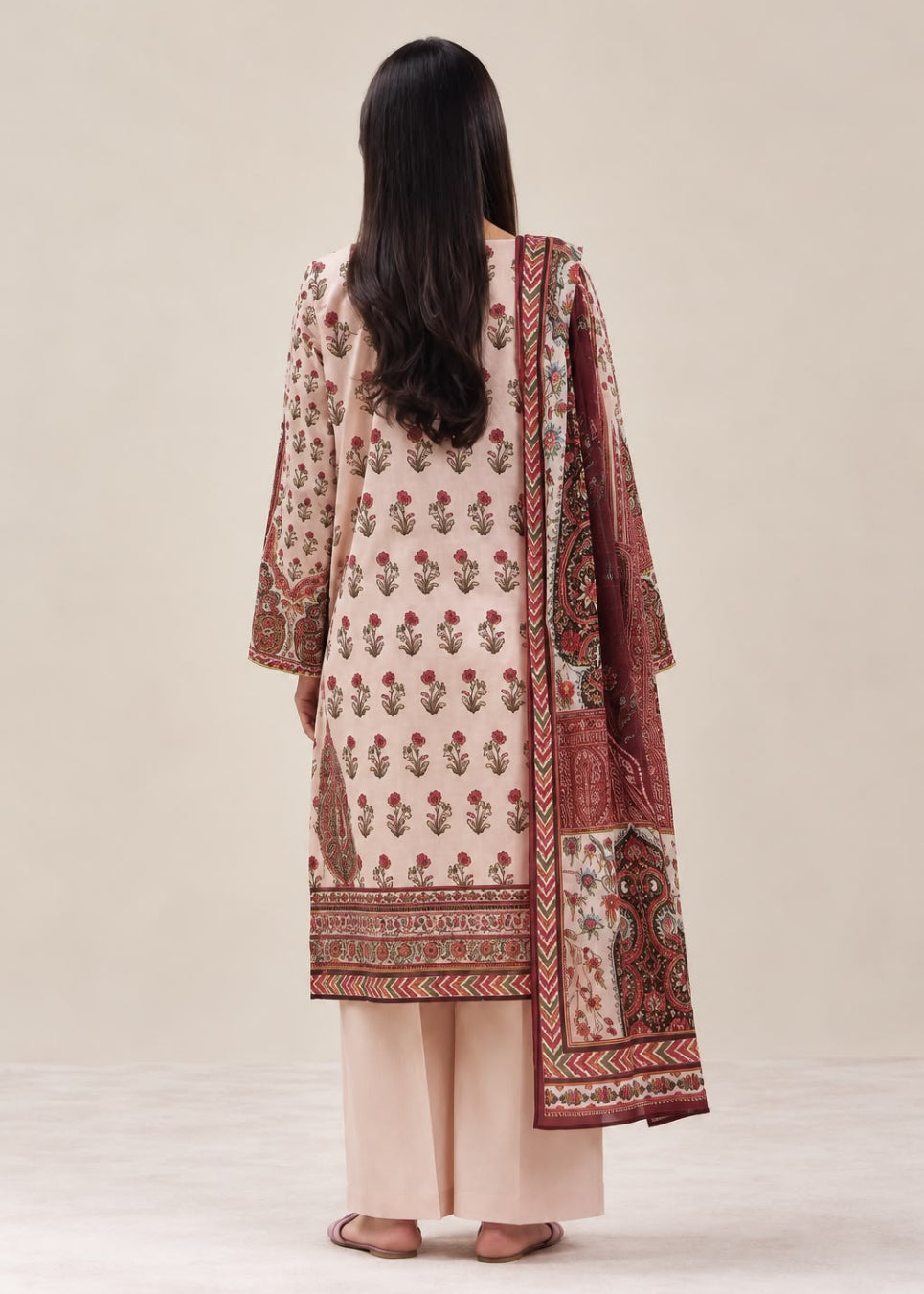 A woman viewed from behind wearing a traditional three-piece salwar kameez suit in beige, featuring a printed kurta with floral motifs, wide sleeves, matching trousers, and a patterned dupatta, set against a solid light beige background.