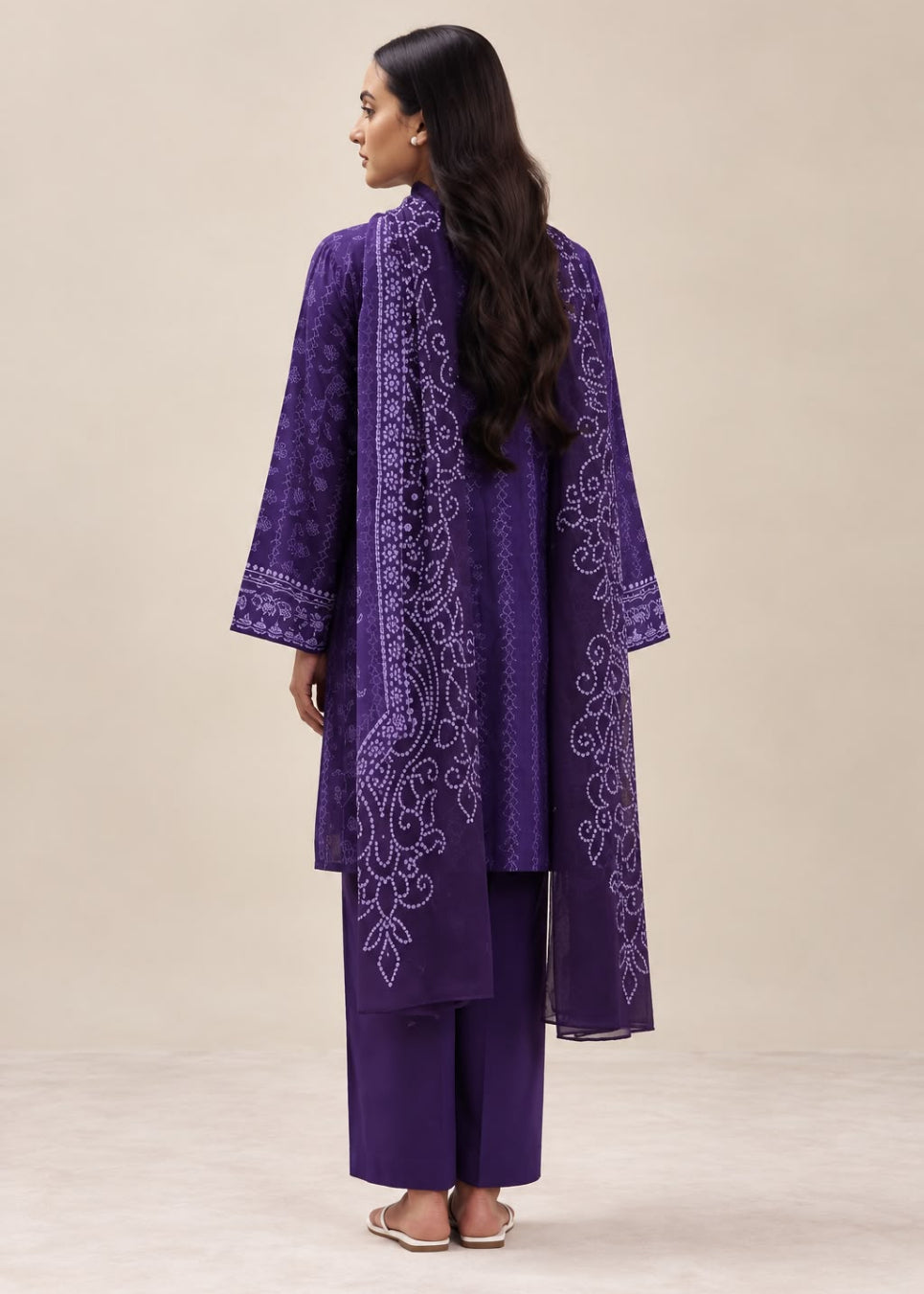 A woman shown from the back wearing a purple traditional outfit with a long embroidered kurta and matching pants, standing against a plain beige studio background.