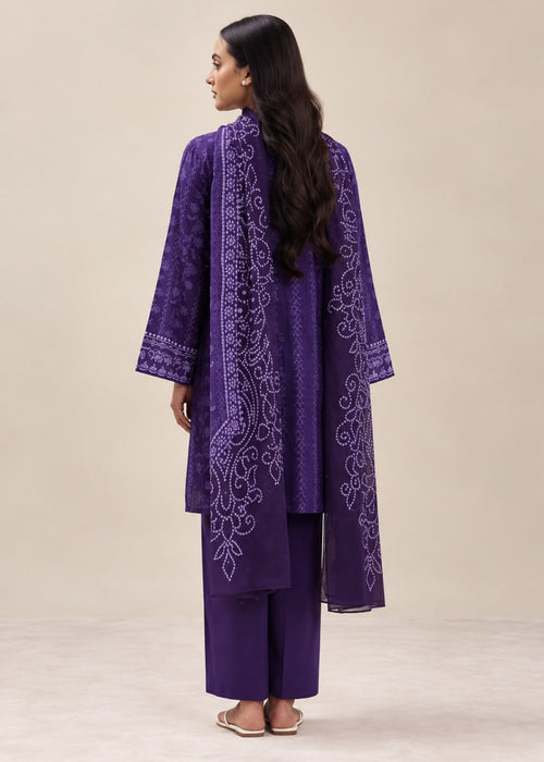 A woman shown from the back wearing a purple traditional outfit with a long embroidered kurta and matching pants, standing against a plain beige studio background.