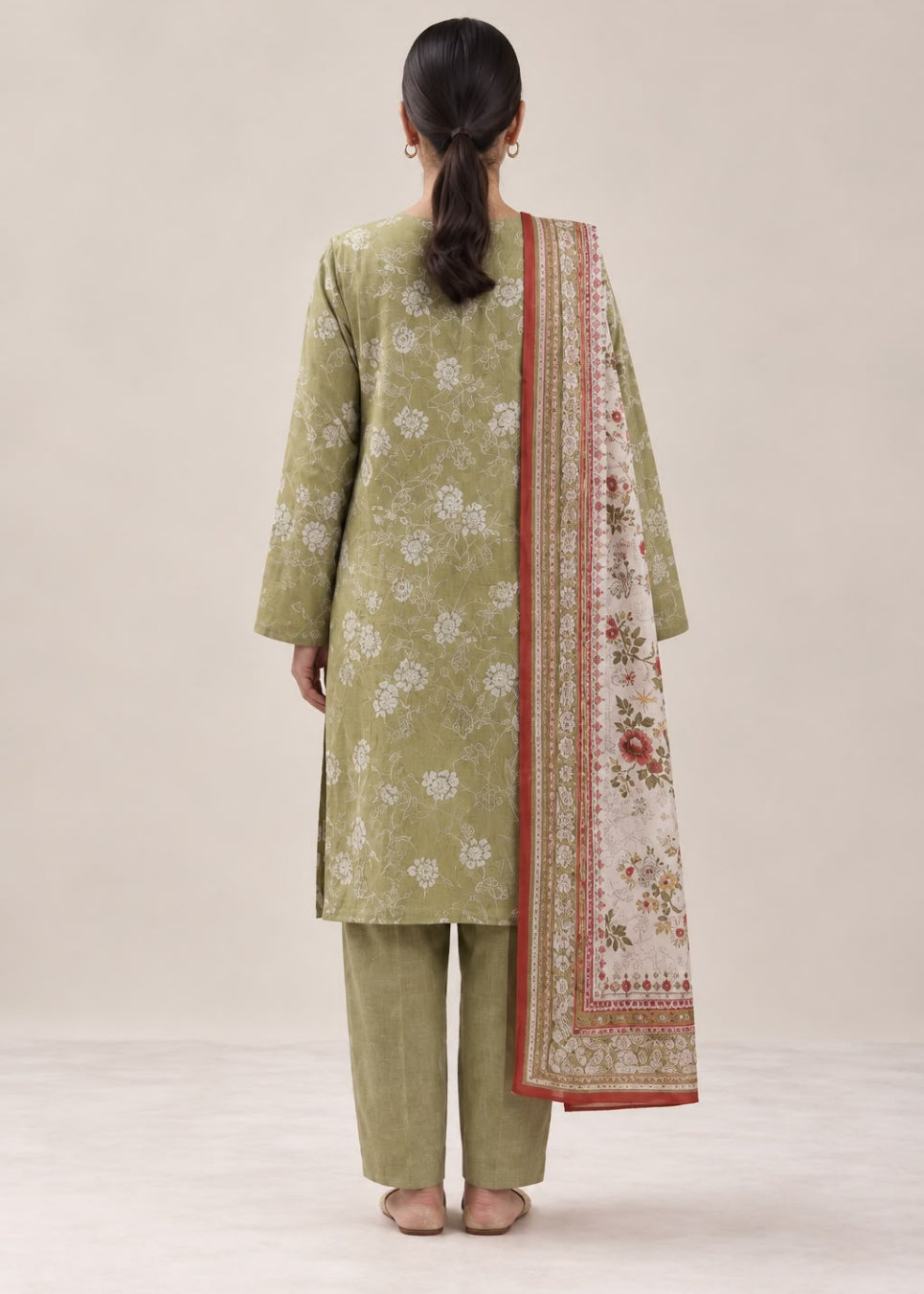 A woman viewed from behind wearing a light olive green three-piece traditional outfit with white floral prints on the kurta and pants, and a cream dupatta with a red border and floral pattern, set against a plain beige studio background.
