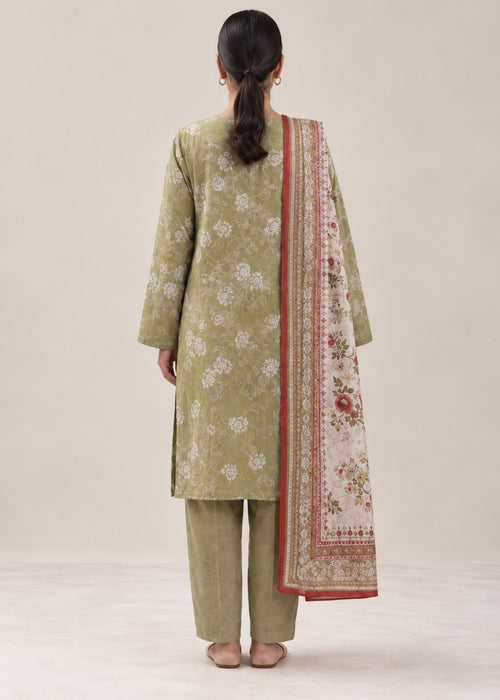 A woman viewed from behind wearing a light olive green three-piece traditional outfit with white floral prints on the kurta and pants, and a cream dupatta with a red border and floral pattern, set against a plain beige studio background.