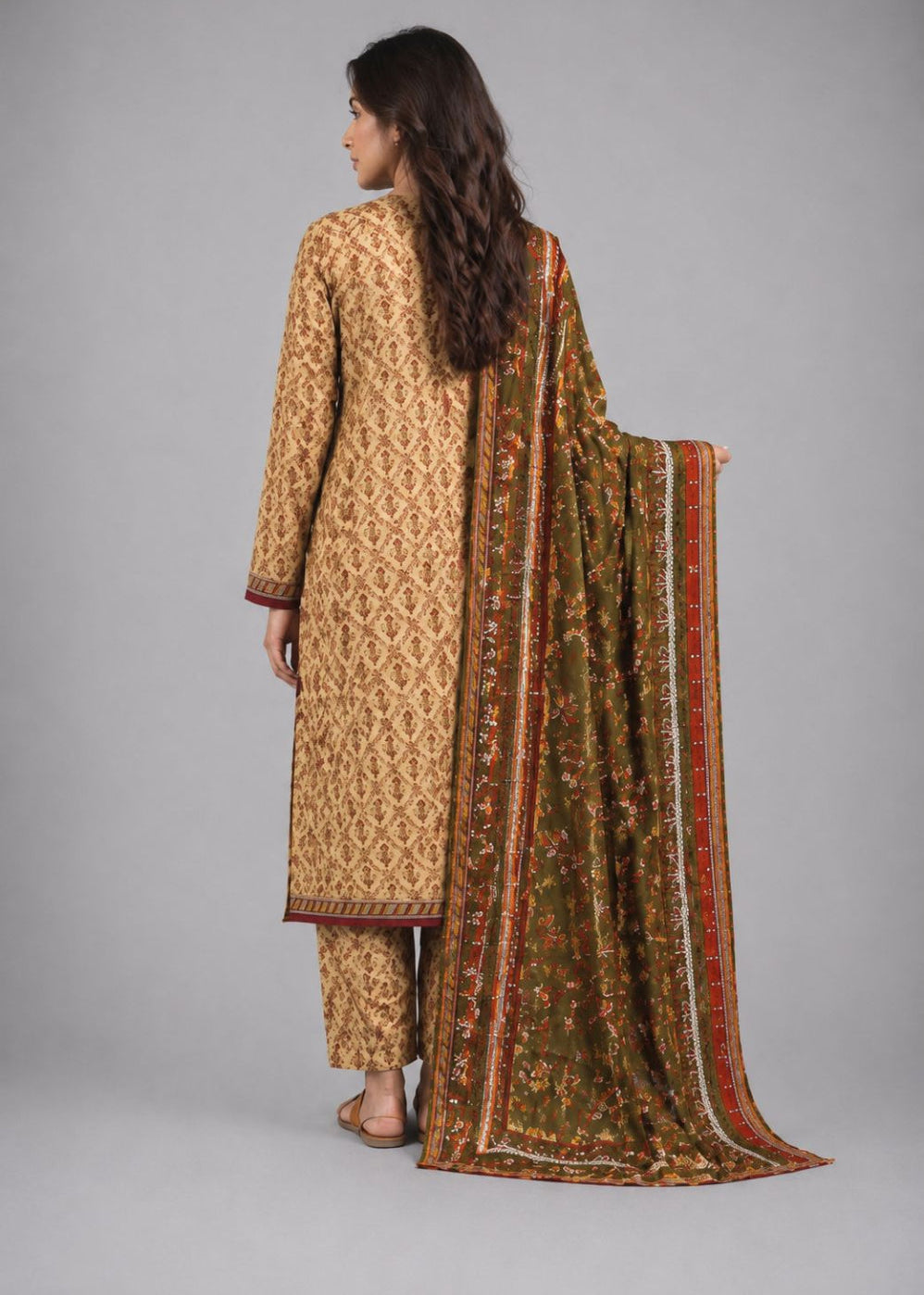 A woman shown from the back wearing a traditional patterned beige kurta with matching pants and a long olive green dupatta featuring red and gold borders, set against a plain gray studio background.