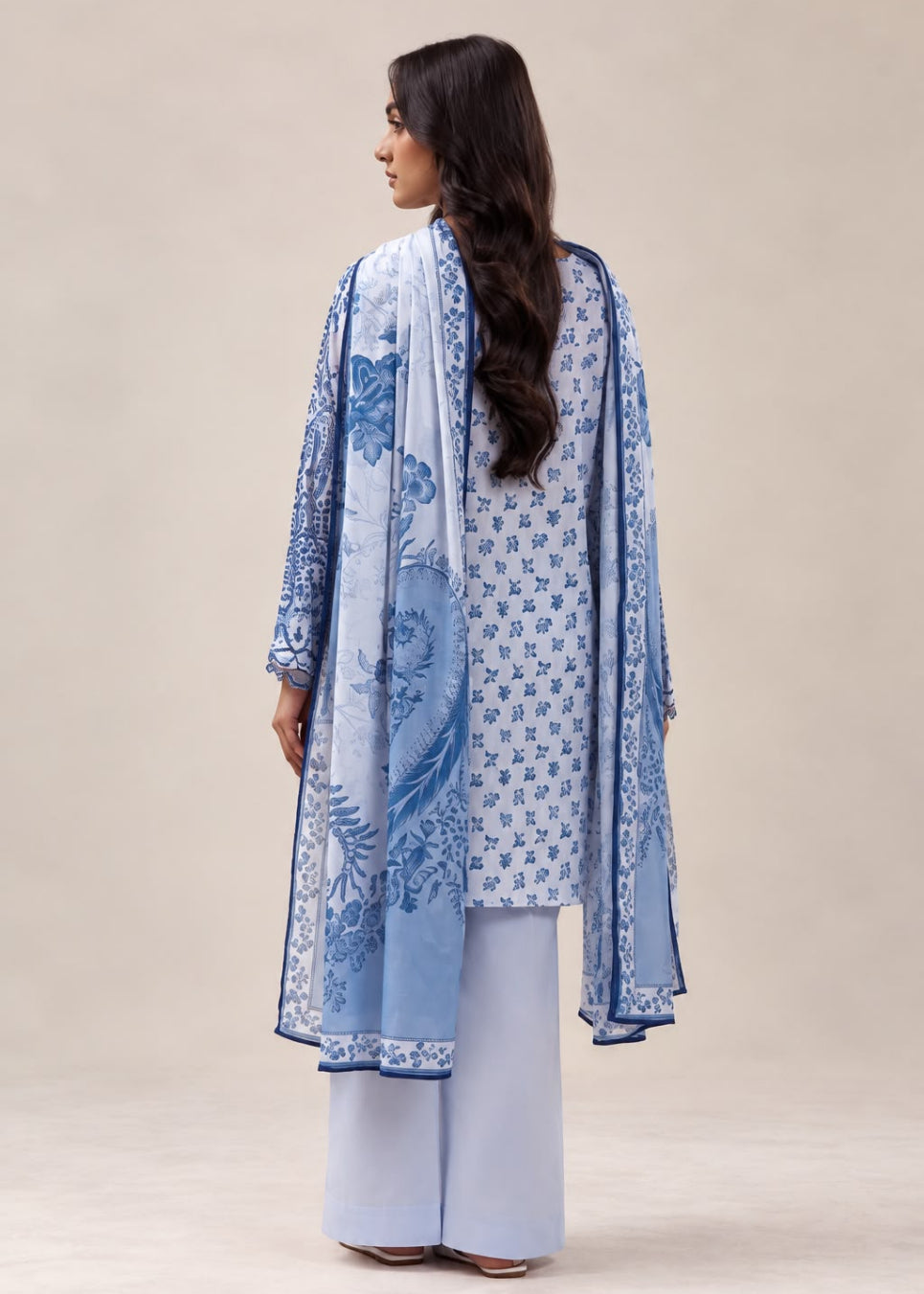 A woman shown from the back wearing a light blue and white traditional outfit with a matching printed dupatta, set against a plain beige studio background.