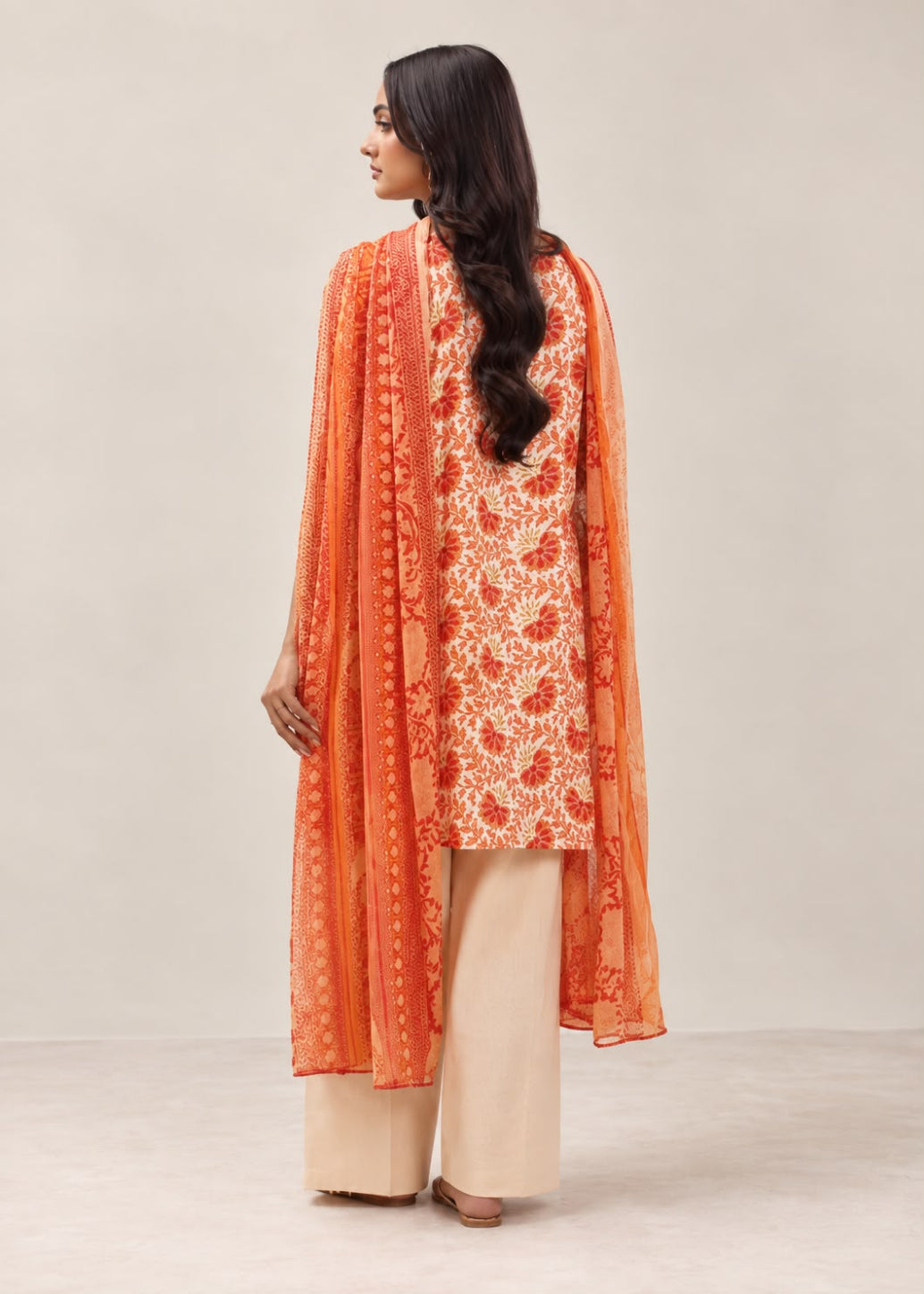 A woman shown from the back wearing a traditional outfit consisting of an orange and beige embroidered kurta, wide-leg beige pants, and a matching sheer orange dupatta, set against a plain light beige studio background.