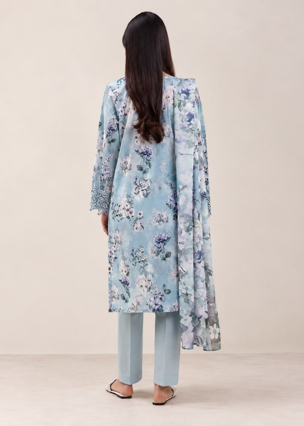 A woman shown from the back wearing a light blue floral kurta with matching light blue pants and black sandals, set against a plain beige studio background.