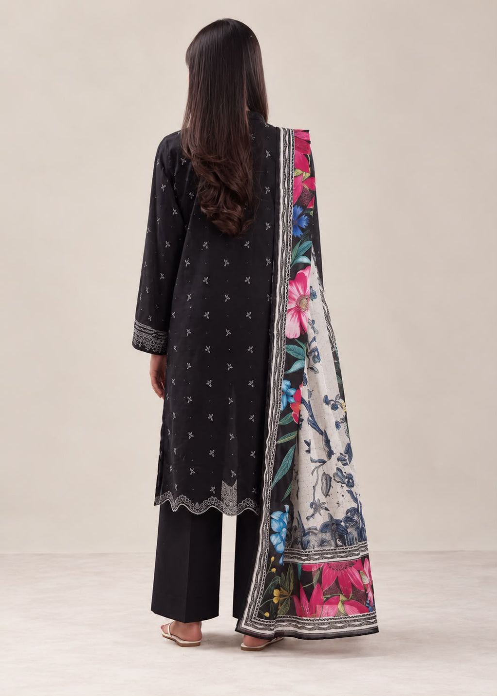 A woman shown from the back wearing a black embroidered kurta with a wide floral printed dupatta, black wide-leg pants, and sandals, set against a plain beige studio background.