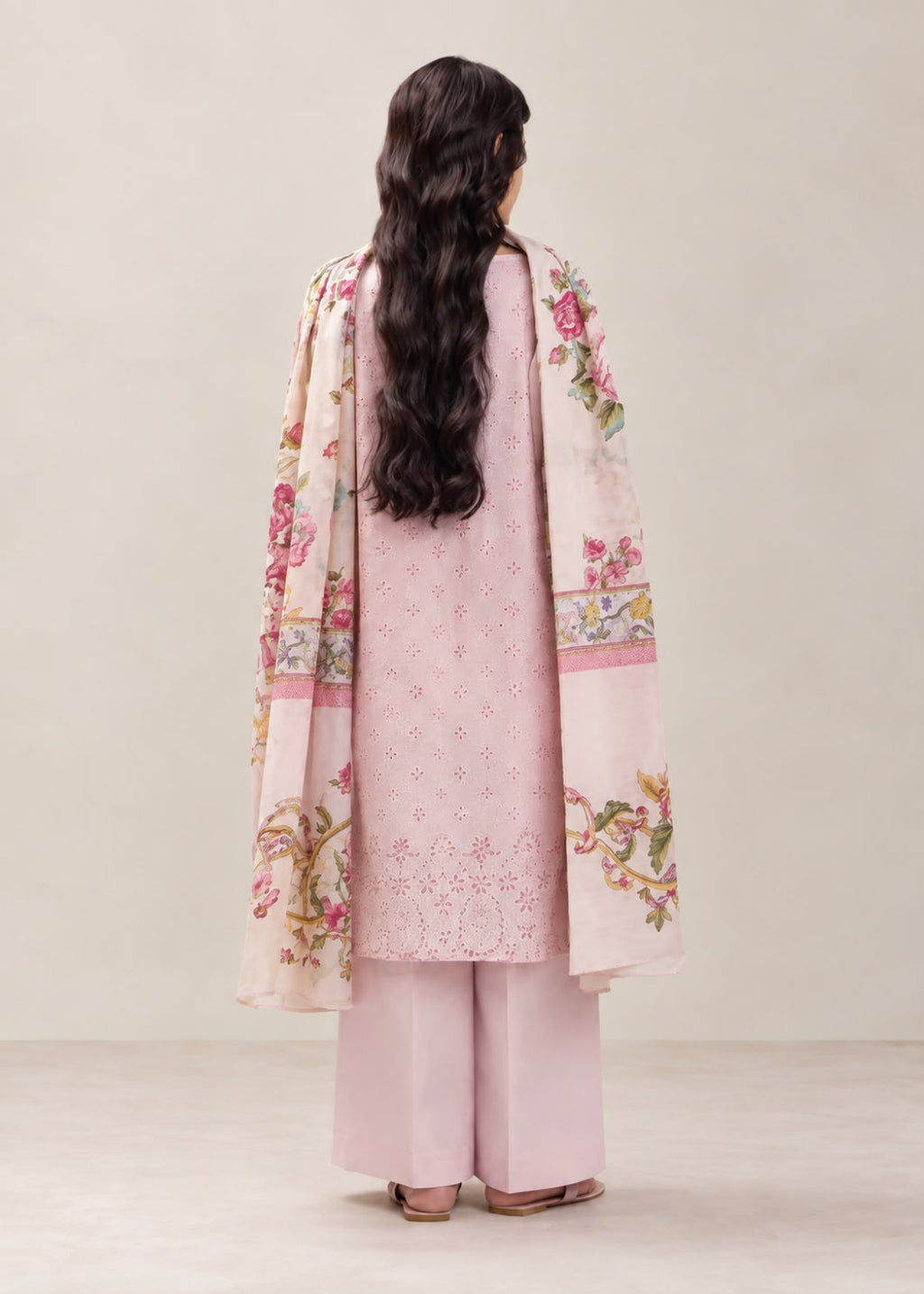 A woman shown from the back wearing a light pink traditional outfit with wide-leg trousers and a matching floral printed outer layer, photographed against a plain warm beige studio background.