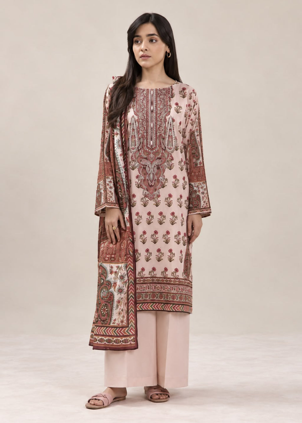 A woman modeling a traditional three-piece salwar kameez suit in dusty pink and brown. The kurta features intricate brown and beige floral and geometric patterns, paired with light pink wide-leg pants and a matching dupatta.