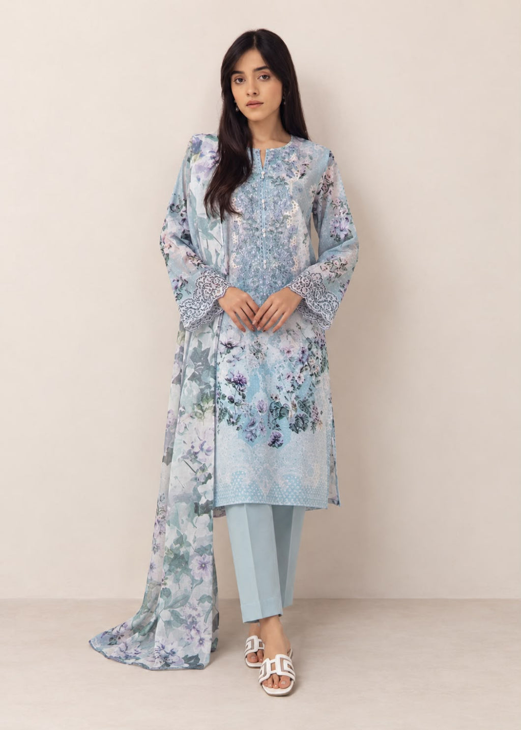 A woman modeling a light blue floral printed traditional outfit with a long kurta, matching pants, and a dupatta, standing against a plain beige studio background.