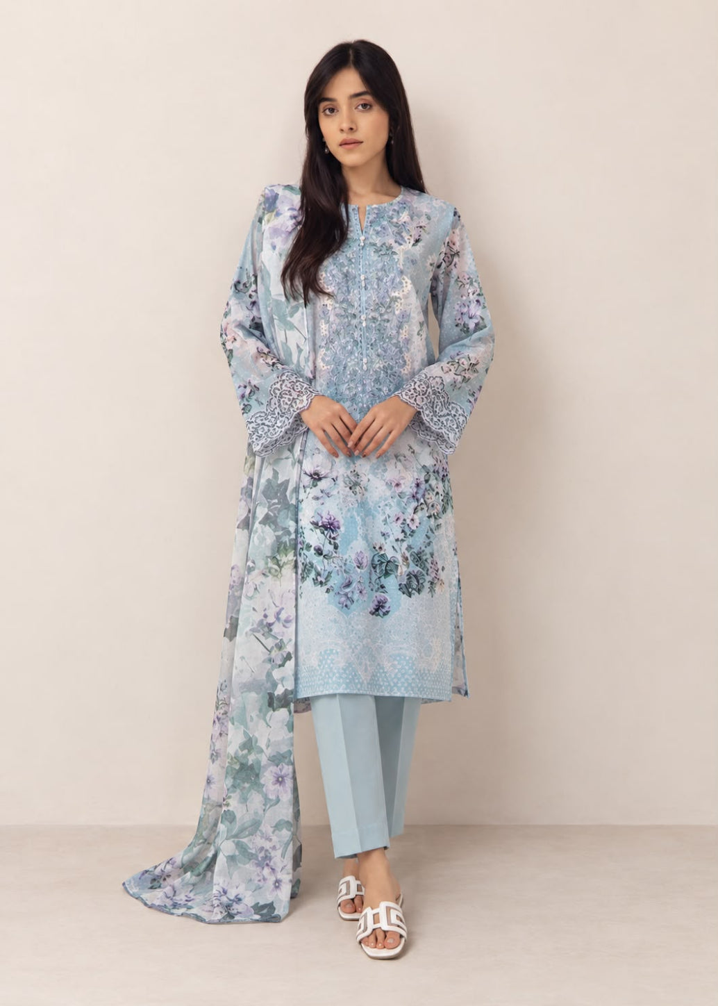 A woman modeling a light blue floral printed traditional outfit with a long kurta, matching pants, and a dupatta, standing against a plain beige studio background.