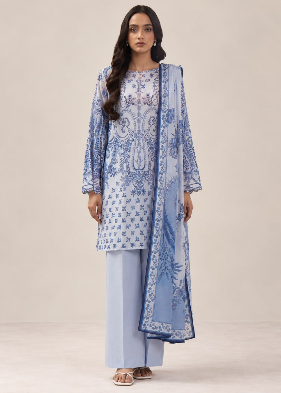 A woman modeling a light blue and white printed traditional outfit with a matching dupatta, consisting of a printed kurta, light blue pants, and a blue patterned scarf, set against a plain beige studio background.