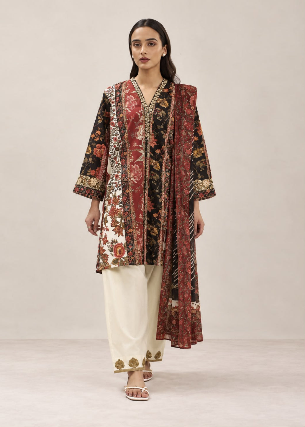 A woman modeling a traditional South Asian outfit featuring a long-sleeved kurta with a complex multicolor floral and geometric print, paired with cream-colored pants and white sandals, set against a plain beige studio background.