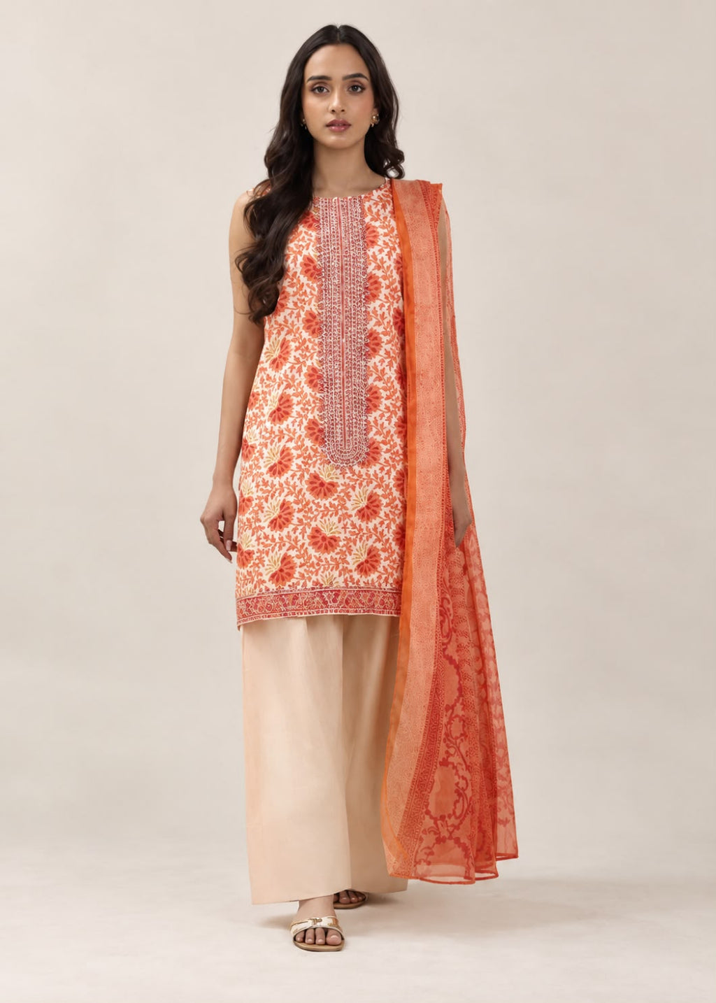 A woman modeling a traditional Indian salwar kameez suit set. The set features a sleeveless cream-colored kurta with orange and red floral embroidery, paired with light beige wide-leg trousers and a matching orange patterned dupatta, set against a plain beige studio background.
