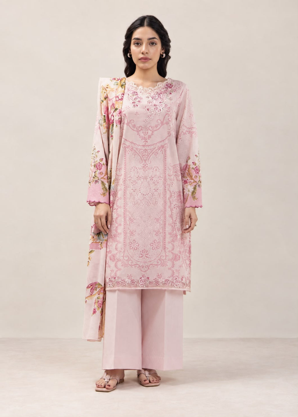 A woman modeling a light pink traditional outfit with floral embroidery and matching wide-leg trousers against a plain beige studio background.