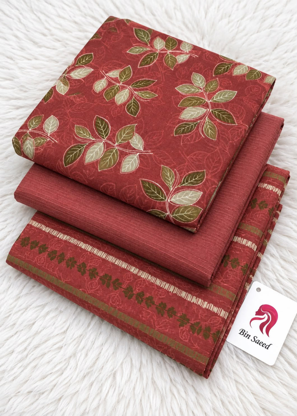 A stack of folded red fabric pieces, likely bed sheets or a bedding set, featuring a green and beige leaf pattern and a decorative border. A white tag with a red horse logo and the text 'Bin Saeed' is attached.