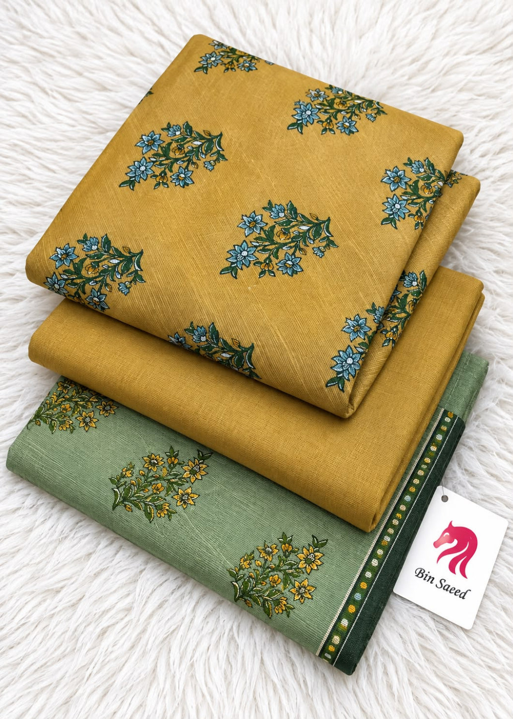 Two folded pieces of printed fabric stacked on a white faux-fur surface. The top piece is mustard yellow with blue floral motifs, and the bottom piece is sage green with yellow floral motifs and a dark green border.