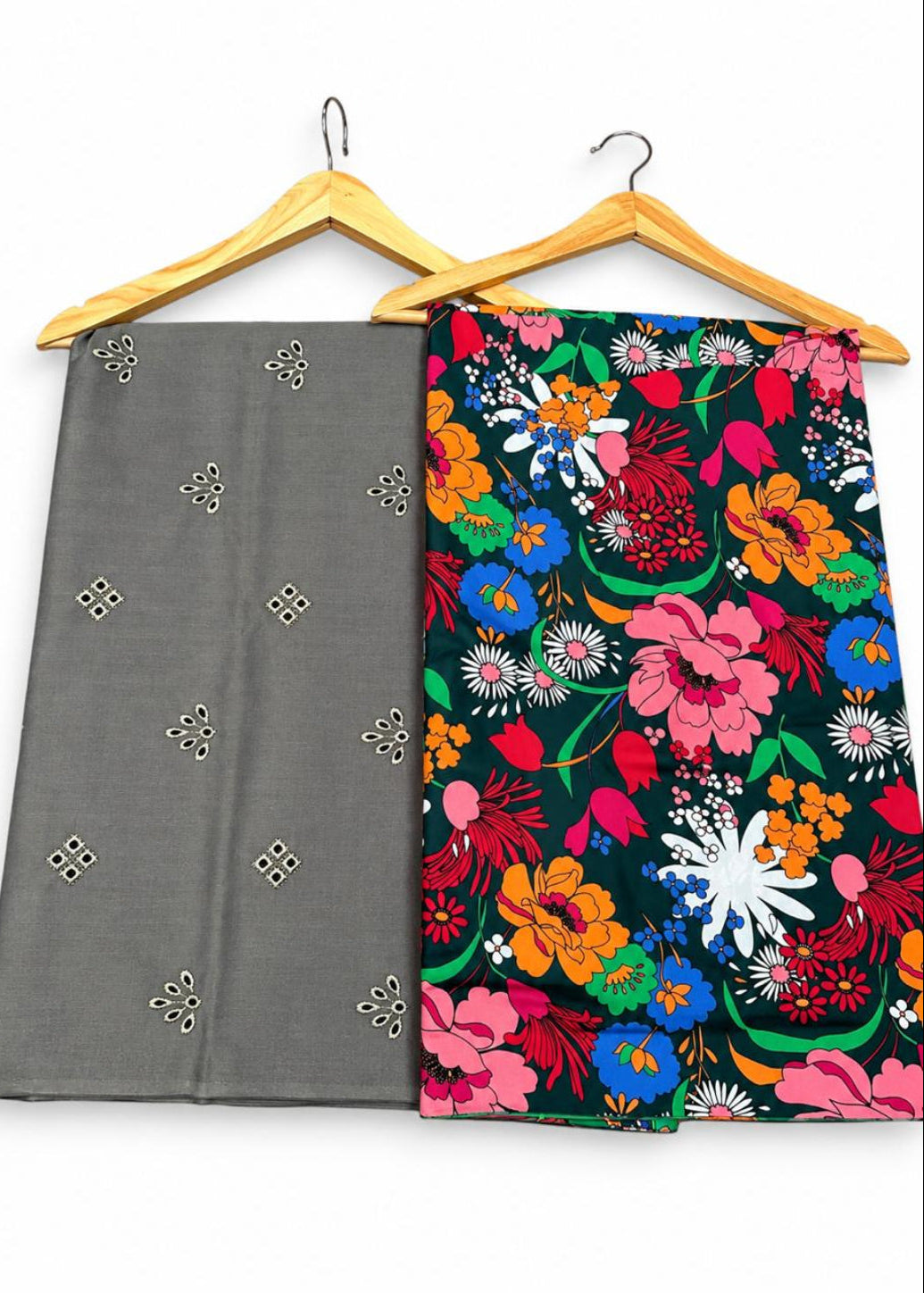 Two folded fabric garments hanging on wooden hangers against a plain white background. The left garment is solid gray with small metallic geometric motifs, and the right garment has a dense multicolor floral print on a dark background with pink, blue, orange, yellow, green, and white flowers.