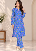 A woman modeling a blue two-piece kurta set with a colorful floral print, standing in a brightly lit indoor setting with a beige wall and a small wooden side table with flowers.