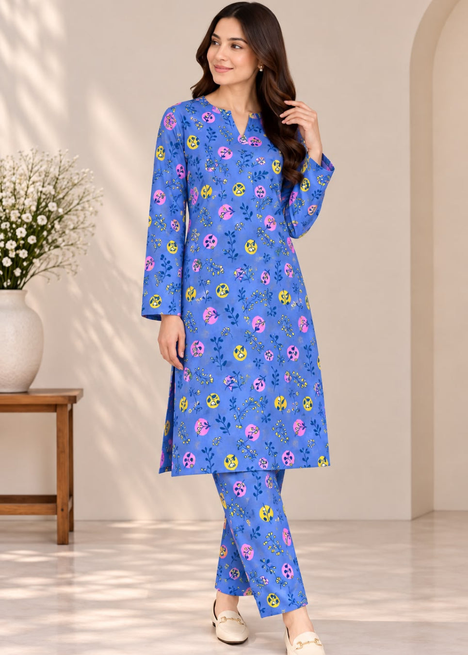 A woman modeling a blue two-piece kurta set with a colorful floral print, standing in a brightly lit indoor setting with a beige wall and a small wooden side table with flowers.