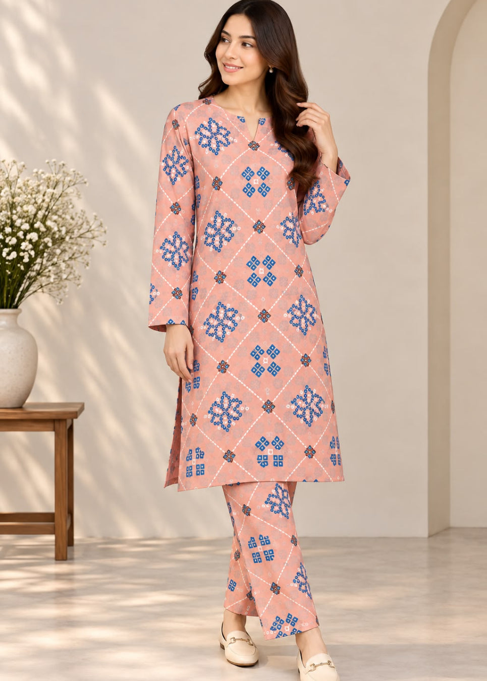 A woman modeling a peach-pink two-piece kurta set with blue and brown geometric patterns, standing in a brightly lit indoor setting with a wooden side table and white flowers on the left.