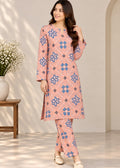 A woman modeling a peach-pink two-piece kurta set with blue and brown geometric patterns, standing in a brightly lit indoor setting with a wooden side table and white flowers on the left.