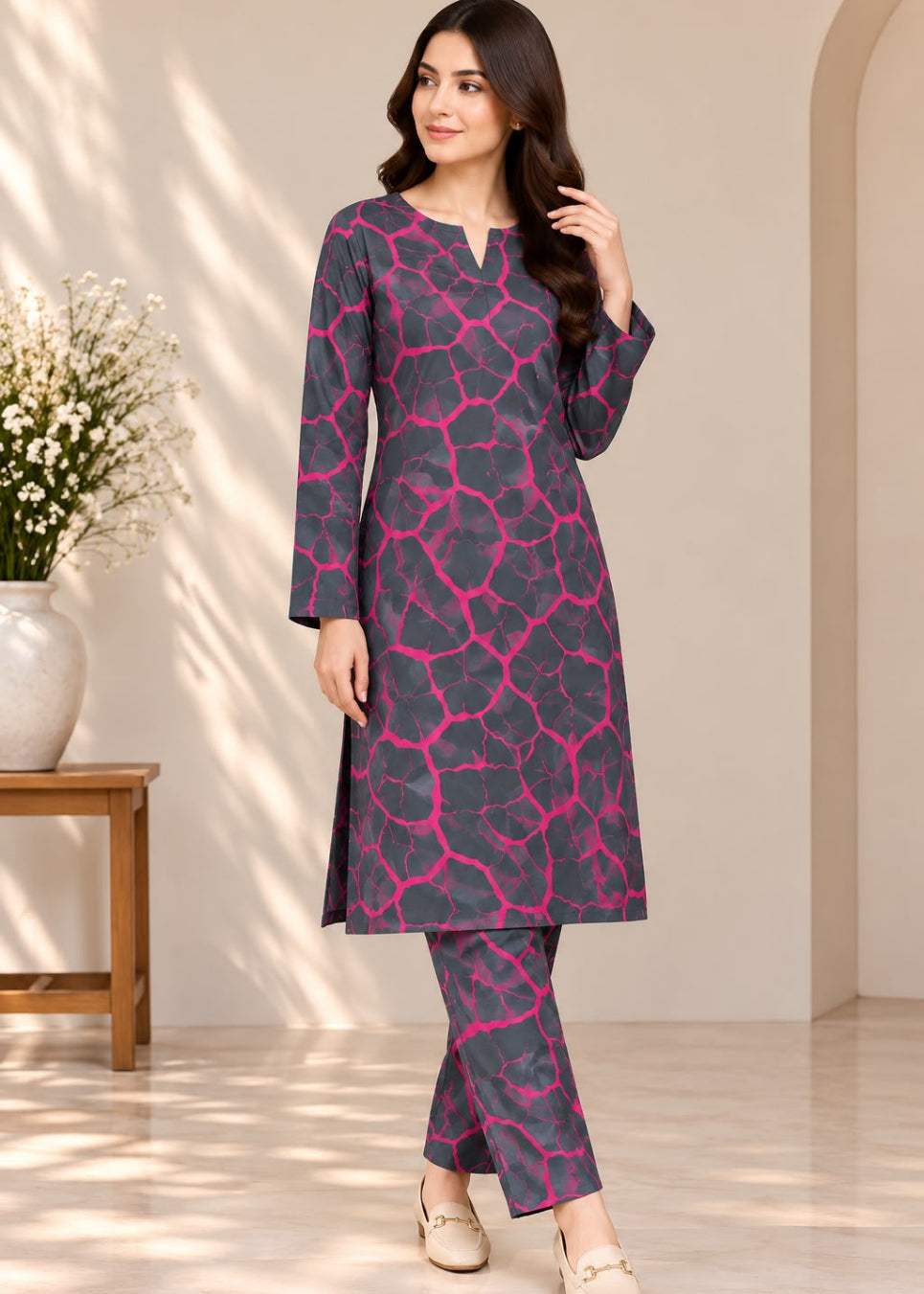 A woman models a three-piece Indian ethnic wear set featuring a dark gray kurta and trousers with a vibrant pink abstract web-like pattern, paired with beige flat shoes in a softly lit indoor setting.