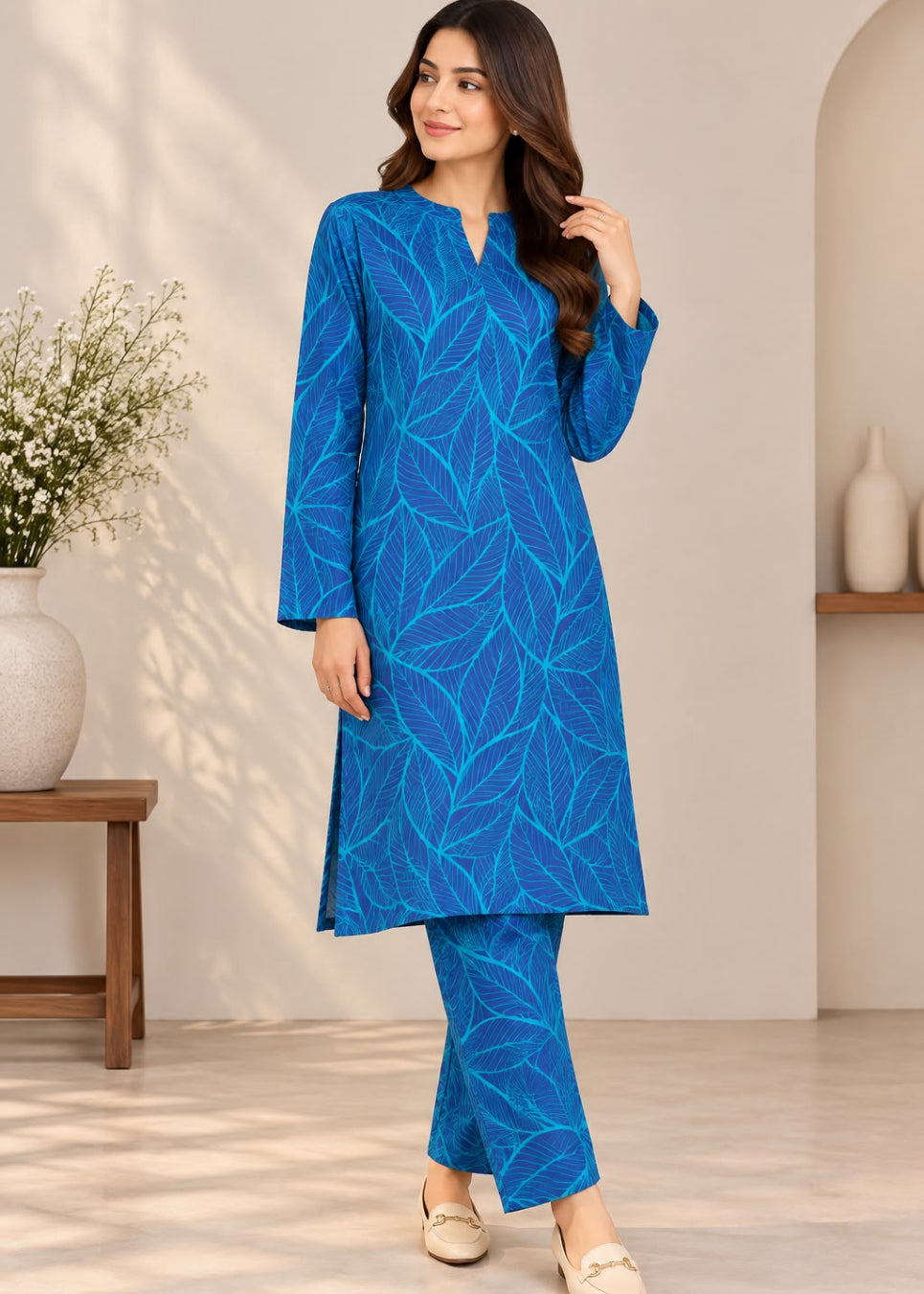 A woman modeling a bright blue three-piece kurta set with a lighter blue leaf pattern, standing in a warm, modern interior setting.