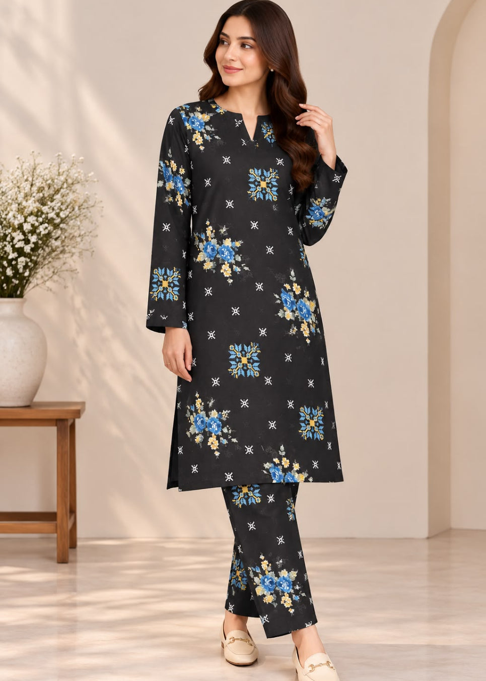 A woman modeling a black long kurta set with a blue and yellow floral print and small white star motifs, paired with matching pants, in a warm beige interior setting.