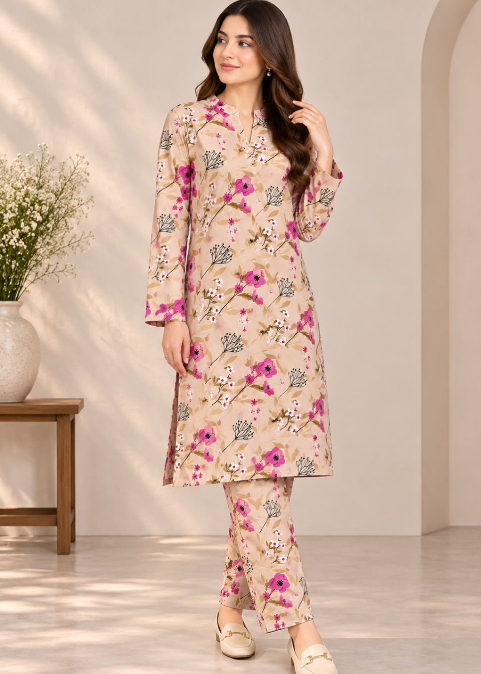 A woman modeling a beige three-piece kurta set with a pink and purple floral print, standing in a brightly lit, minimalist room with neutral beige walls and arches.