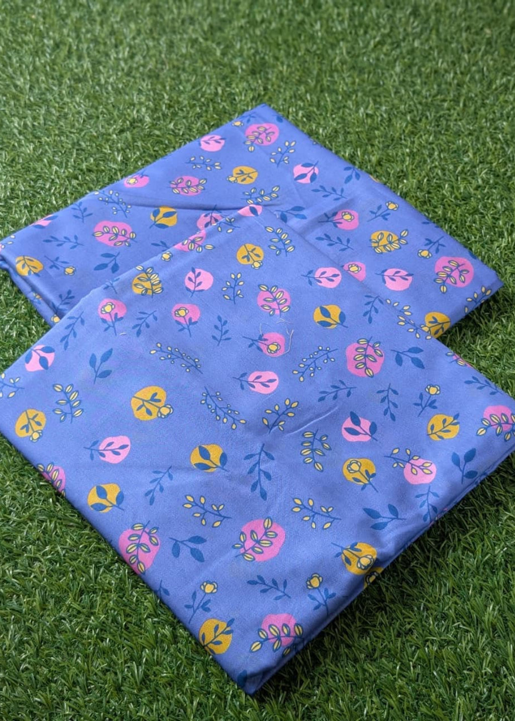 A folded blue fabric item, likely a quilt or blanket, featuring a colorful floral and leaf pattern in pink, yellow, and blue, displayed on green artificial grass.