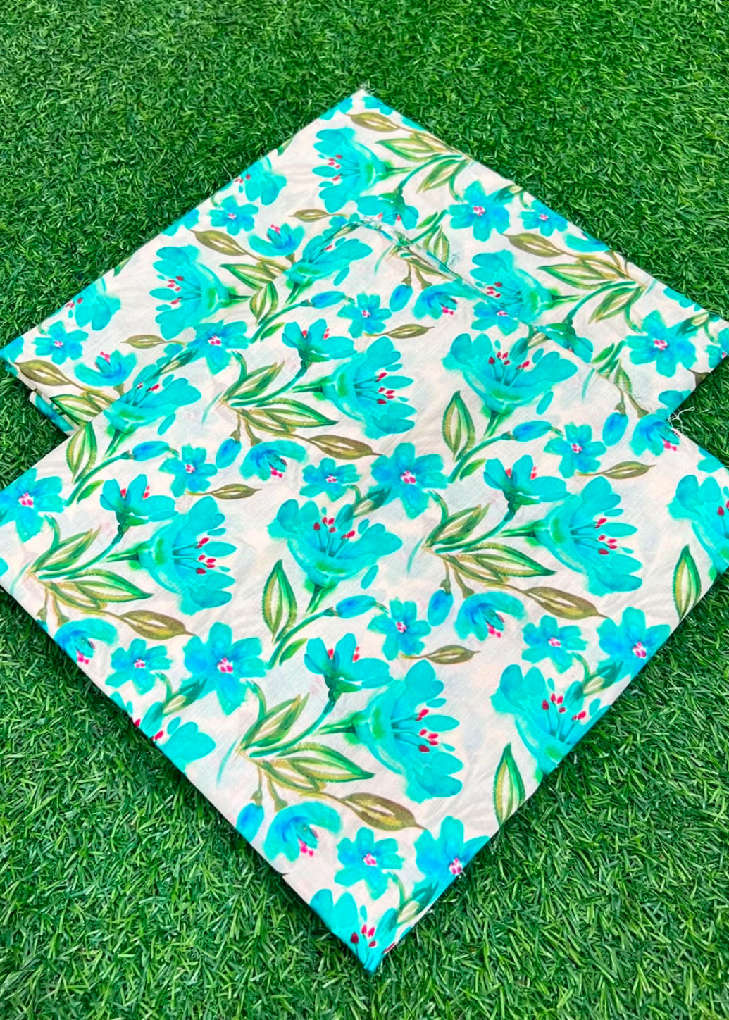 Two square fabric items with a bright blue and green floral print on a white background, laid flat on green artificial grass.